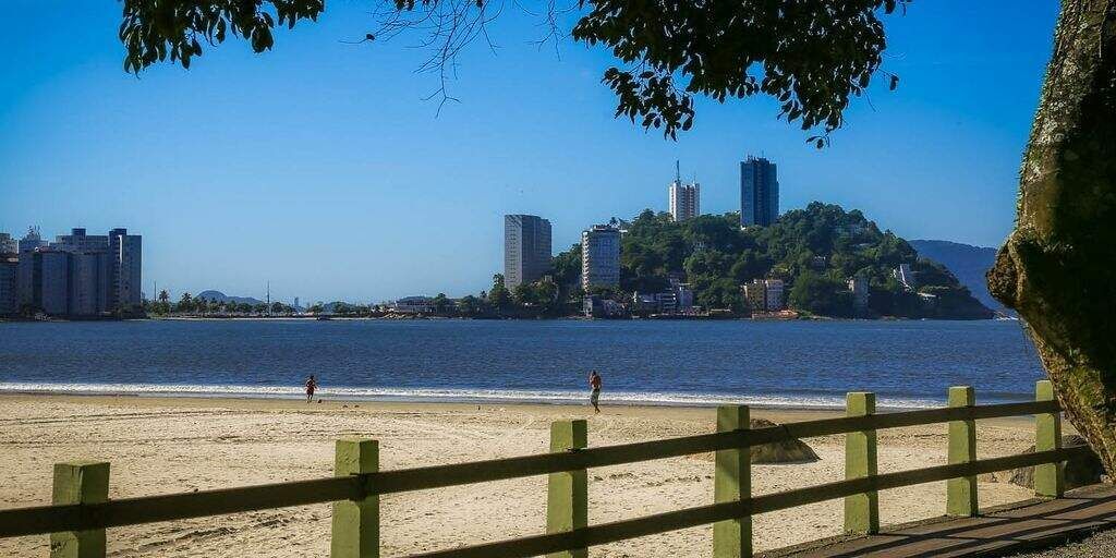 Praia de São Vicente com faixa de areia, oceano e edifícios ao fundo sob um céu azul.