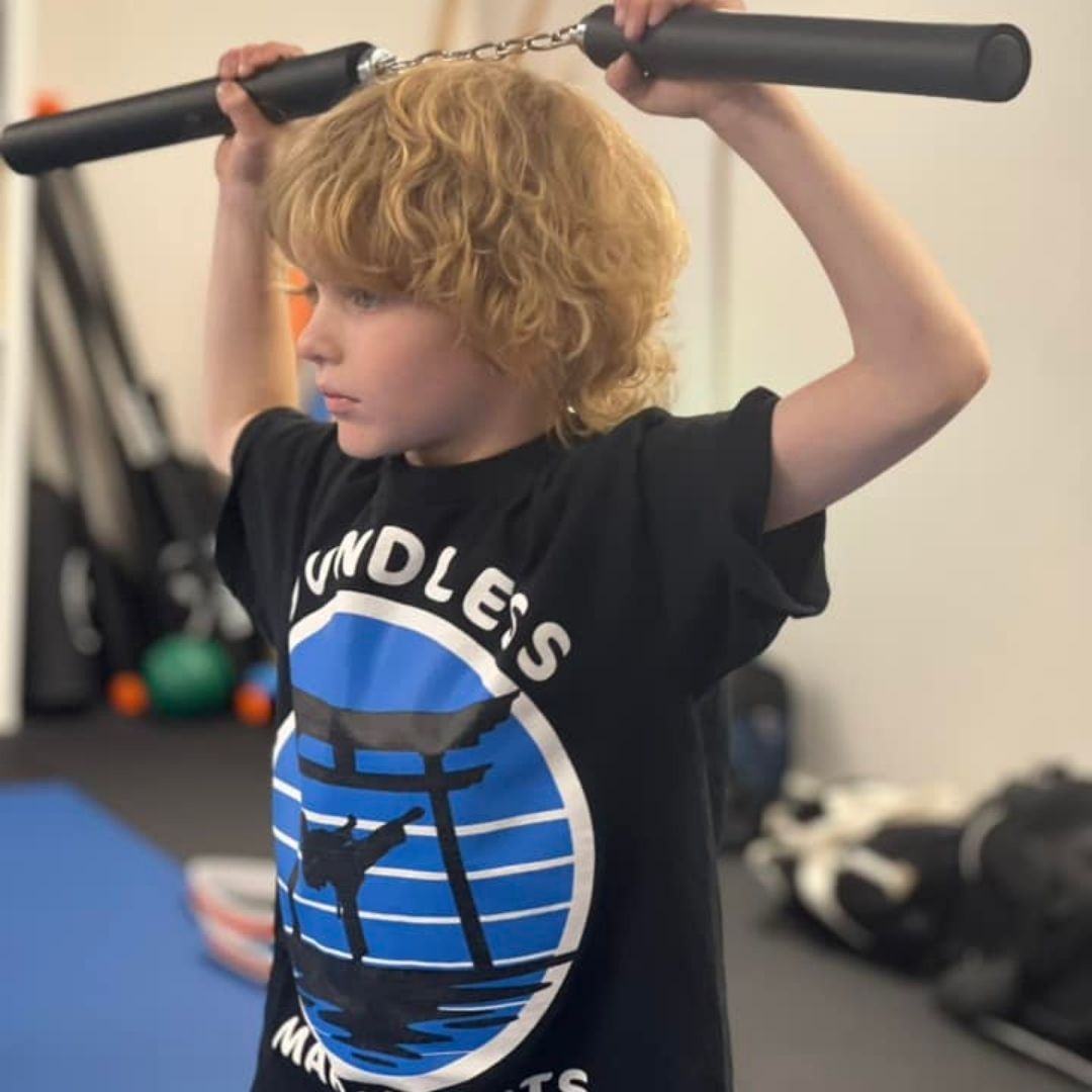 Two children in black and white martial arts uniforms practice a chokehold on a mat. One child smiles.