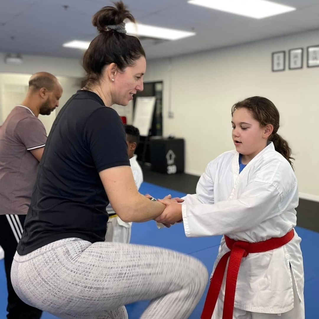 Two children practicing Brazilian Jiu-Jitsu on a mat. One child in white gi has the other in a leg lock. A girl watches.