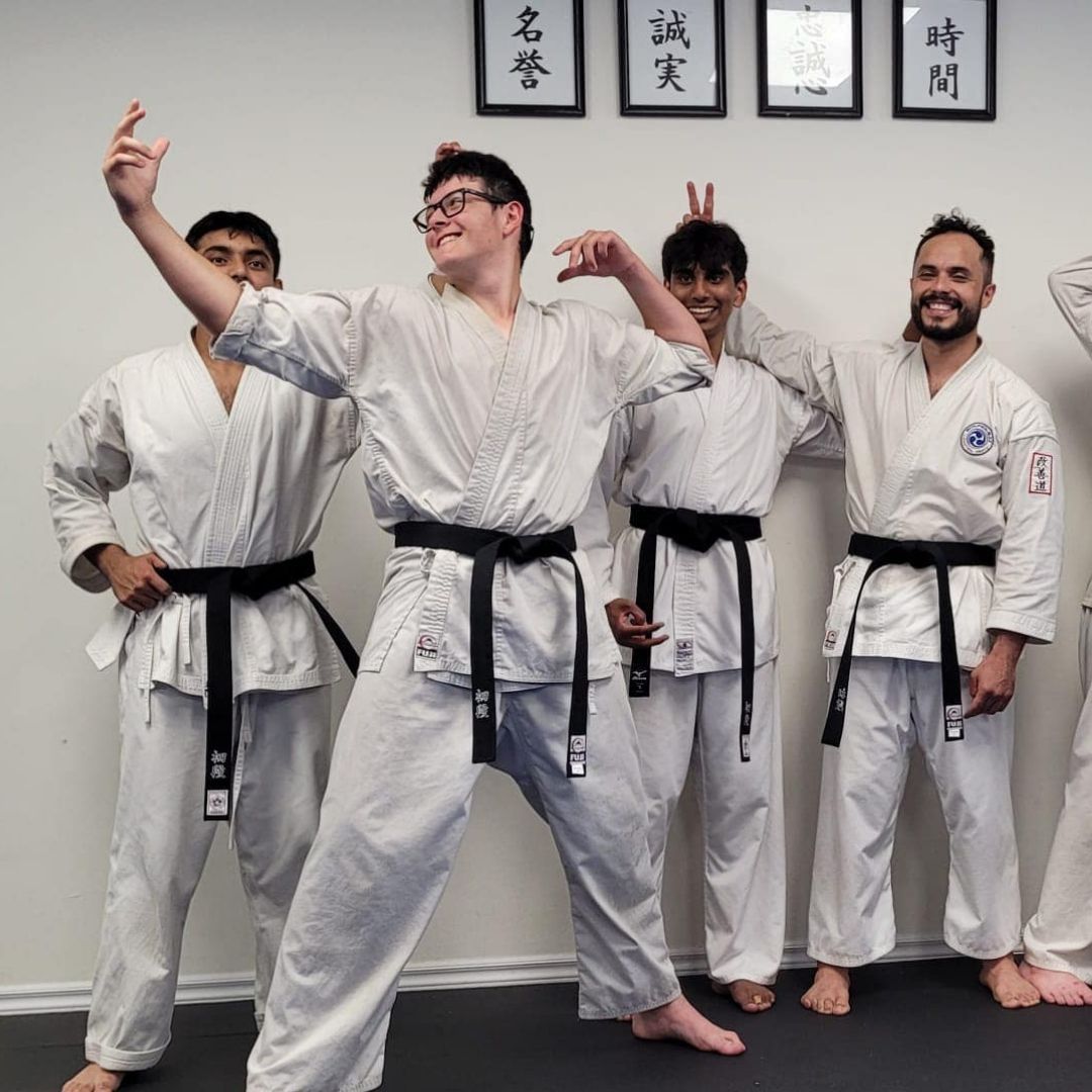 Two men in grappling position on a mat. One embraces the other from behind, both smiling. Gym setting.