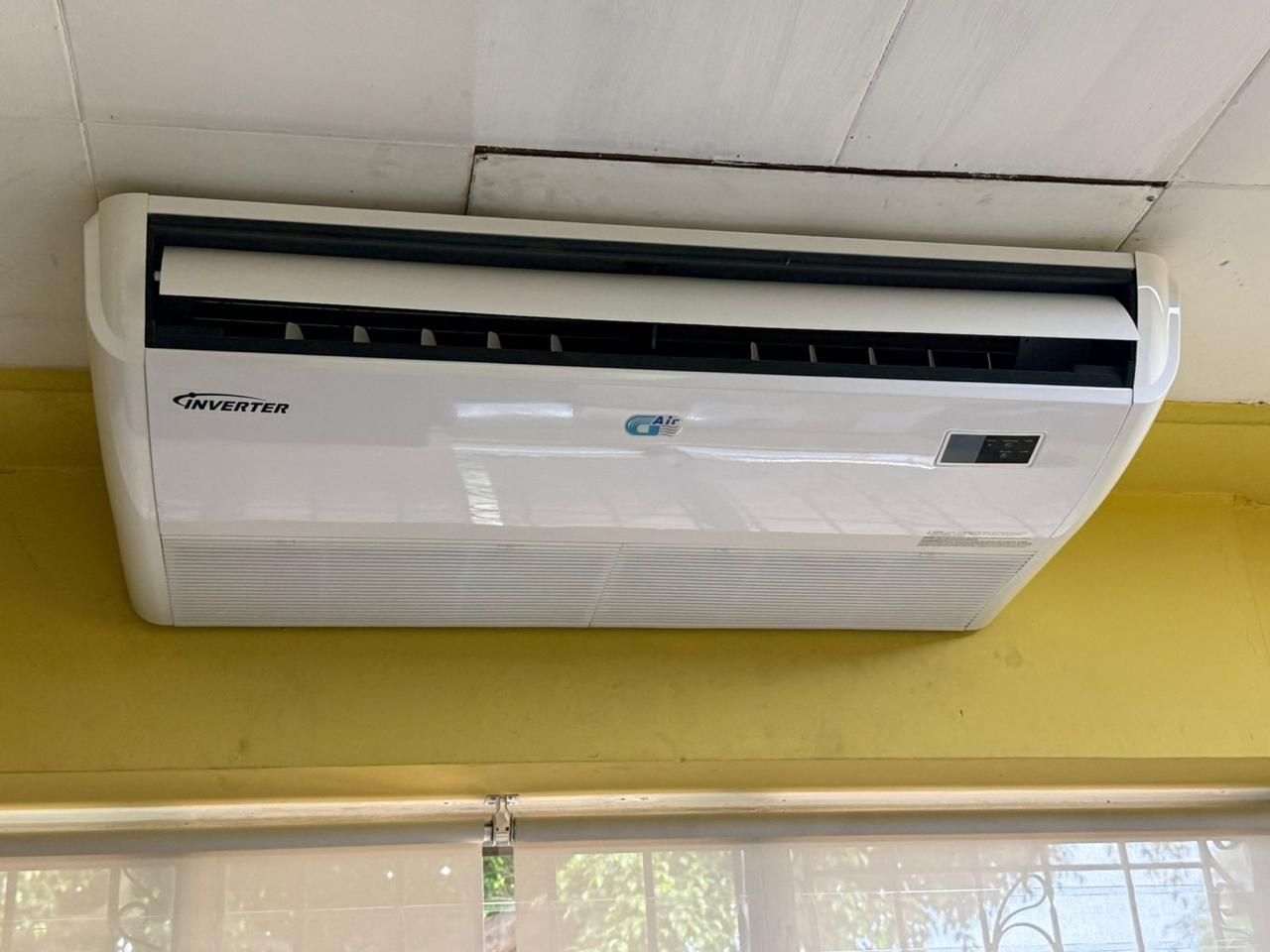 Aire acondicionado split blanco montado en la pared, encima de una ventana con pared y techo amarillos.
