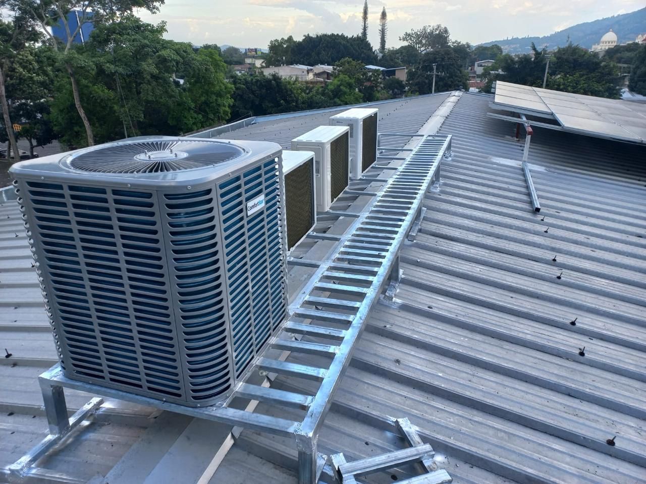 Unidades de aire acondicionado en la azotea y conductos metálicos sobre un techo ondulado al anochecer.