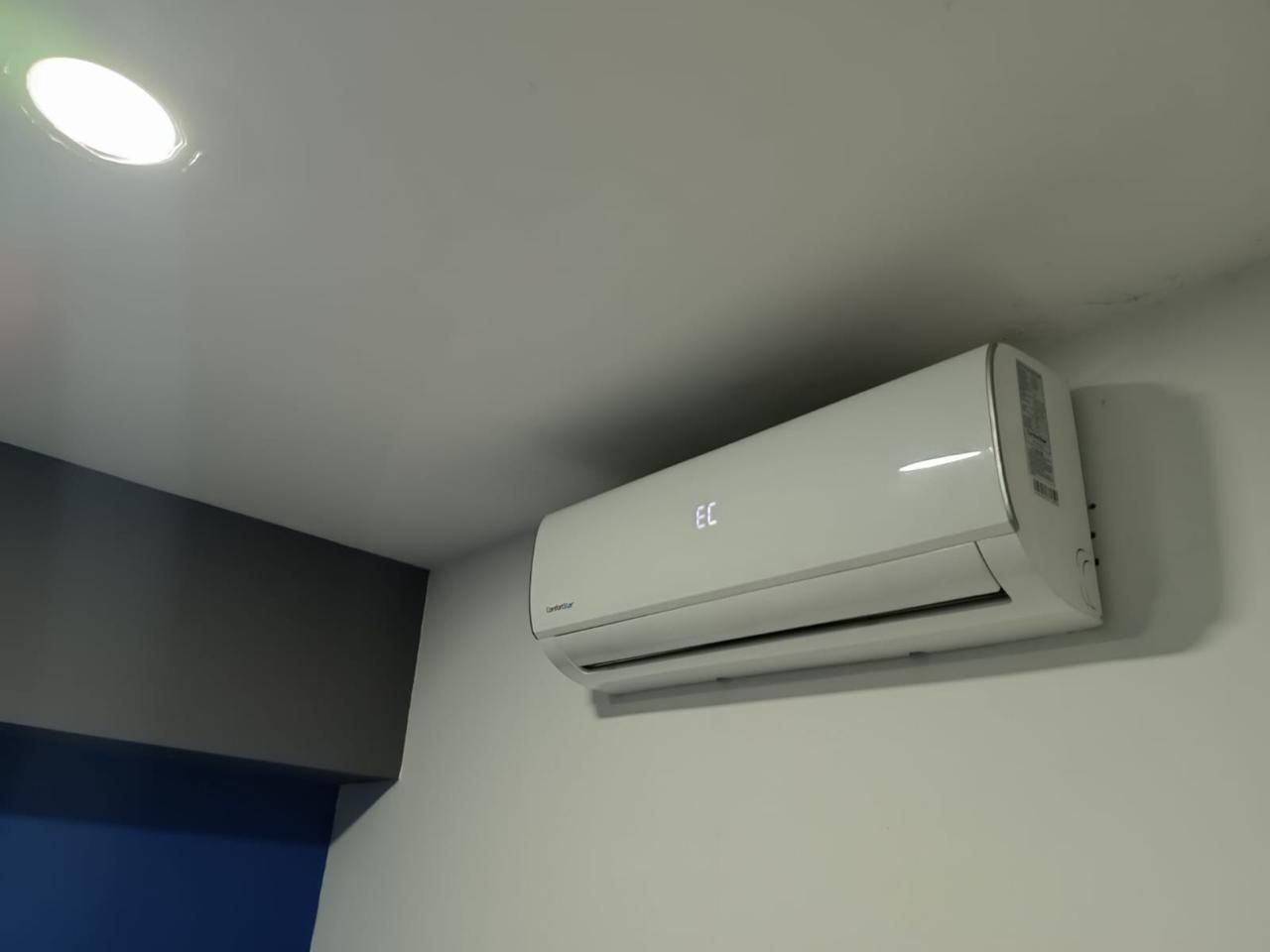 Aire acondicionado blanco montado en la pared sobre una pared gris cerca de una lámpara de techo.