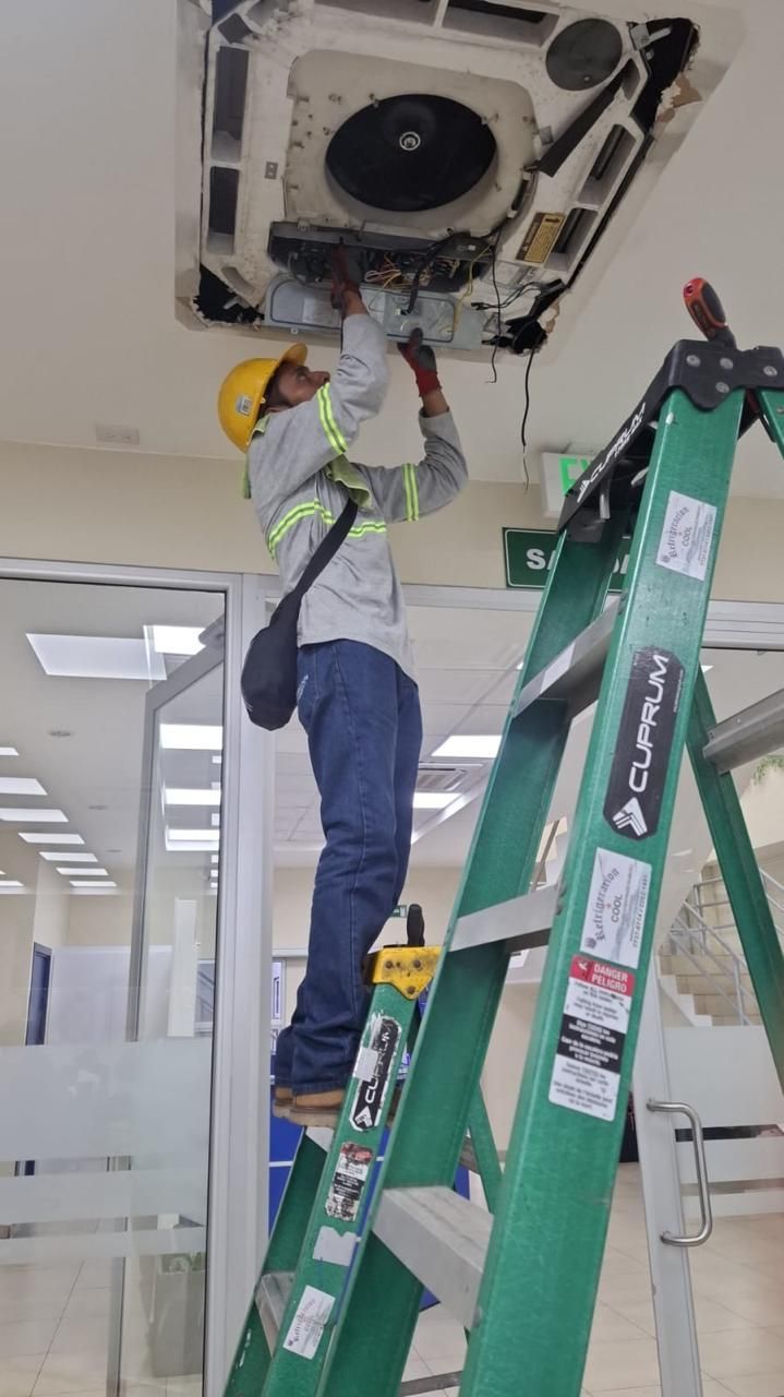 Un trabajador, subido a una escalera verde, repara una unidad de climatización instalada en el techo de una oficina.