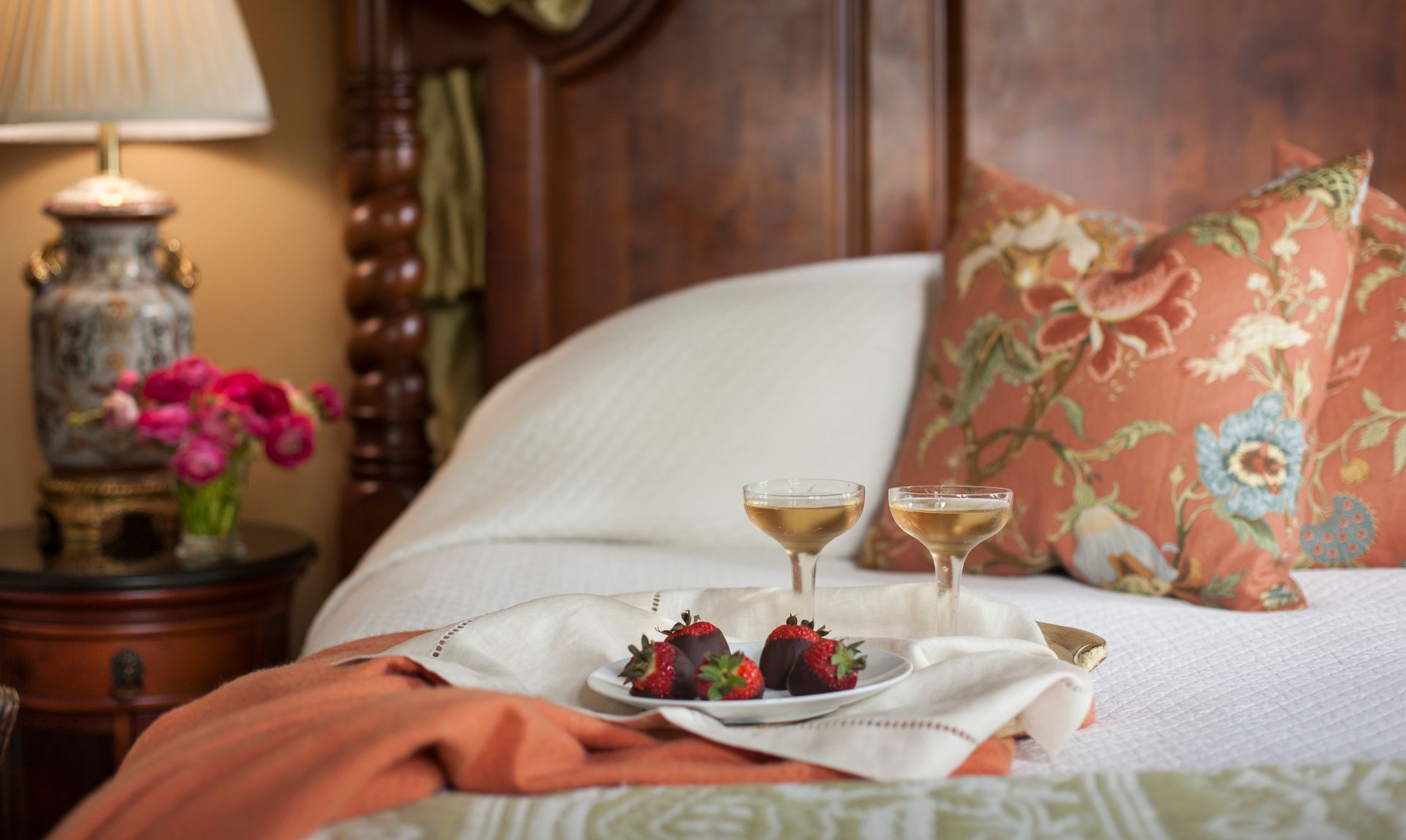 A bedroom with a tray of chocolate-covered strawberries and champagne glasses on a bed with decorative pillows.