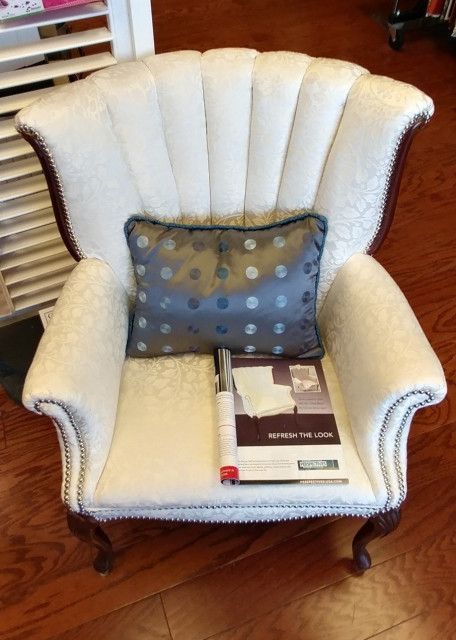 Cream-colored antique armchair with blue pillow and reading material on a wooden floor.