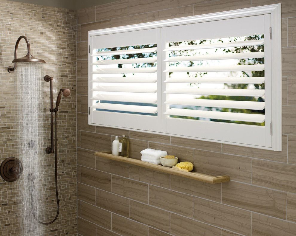 Shower with bronze fixtures and white shuttered window; tan tile.