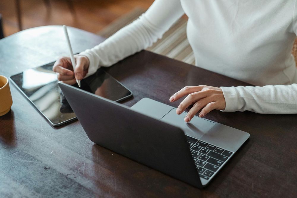 A woman is sitting at a table using a laptop and a tablet.