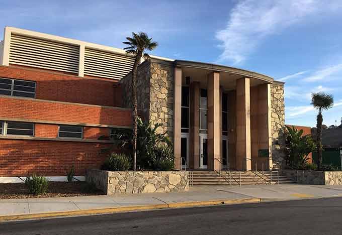 Commercial building with tan and white facade, windows, and parking spaces. Blue sky and greenery.