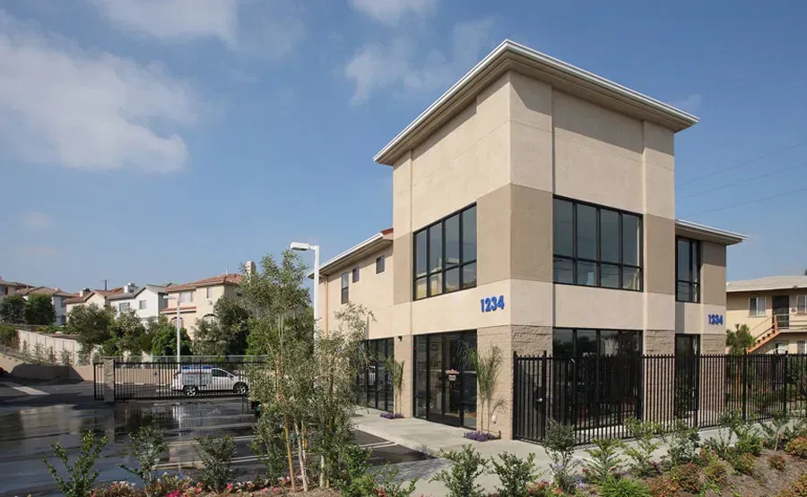 Shopping center with various storefronts under a clear blue sky, cars parked in front.