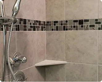 Bathroom shower corner with chrome handheld showerhead, gray tile, mosaic border, and corner shelf