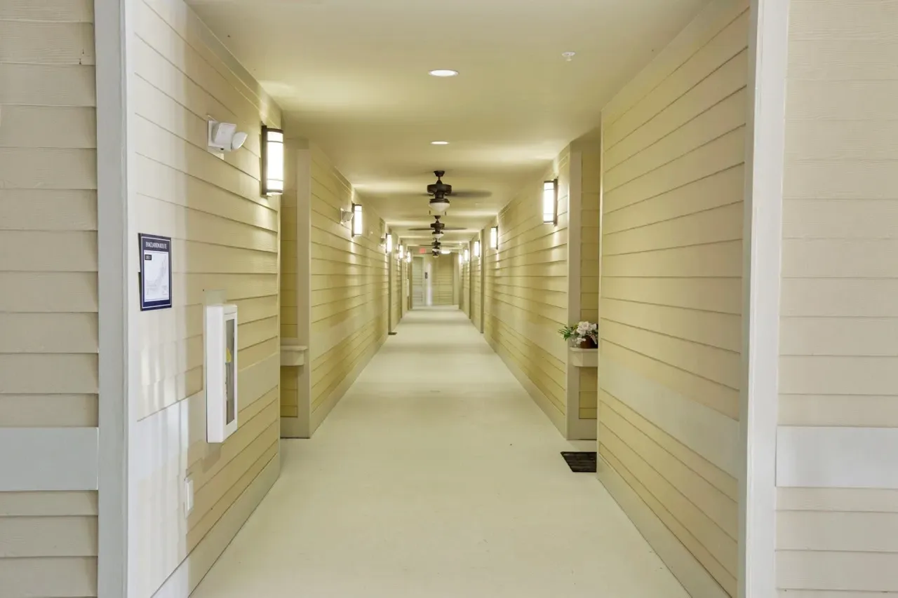 Long interior hallway in an apartment building with beige siding, doors along both walls, and ceiling lights.