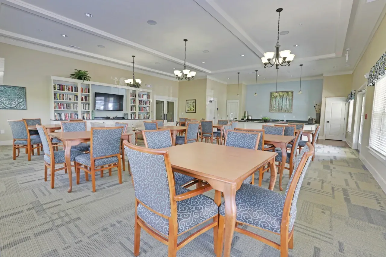 Community lounge with tables, blue patterned chairs, bookshelves, and a wall-mounted TV.