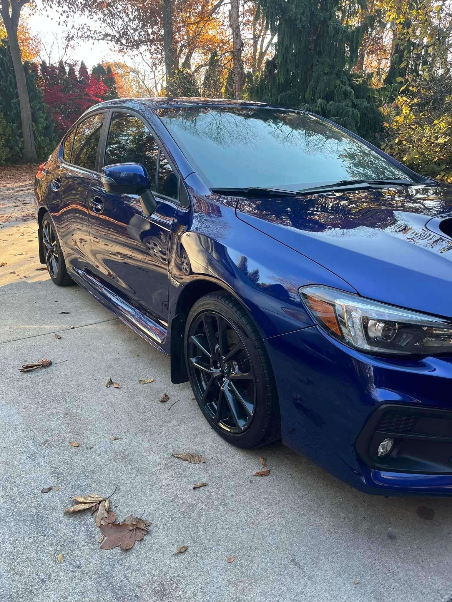 A dark blue Subaru sedan parked on a concrete driveway with autumn leaves scattered on the ground.