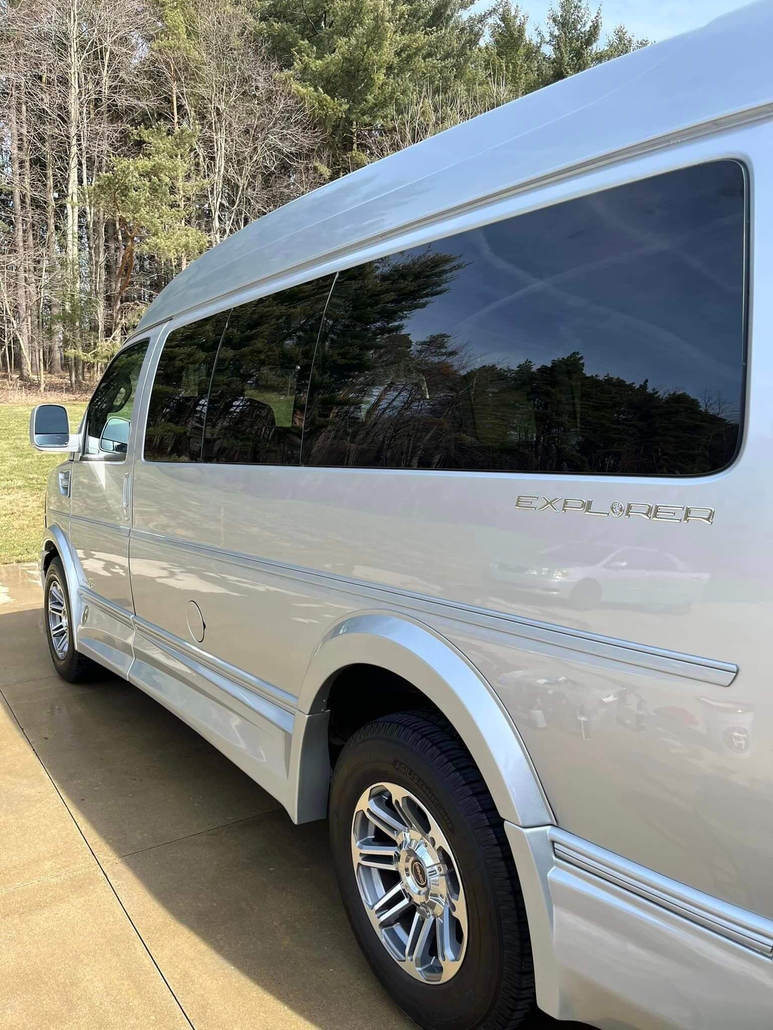 A silver conversion van parked outdoors on a paved surface, featuring a high roof, tinted windows, and chrome wheels.