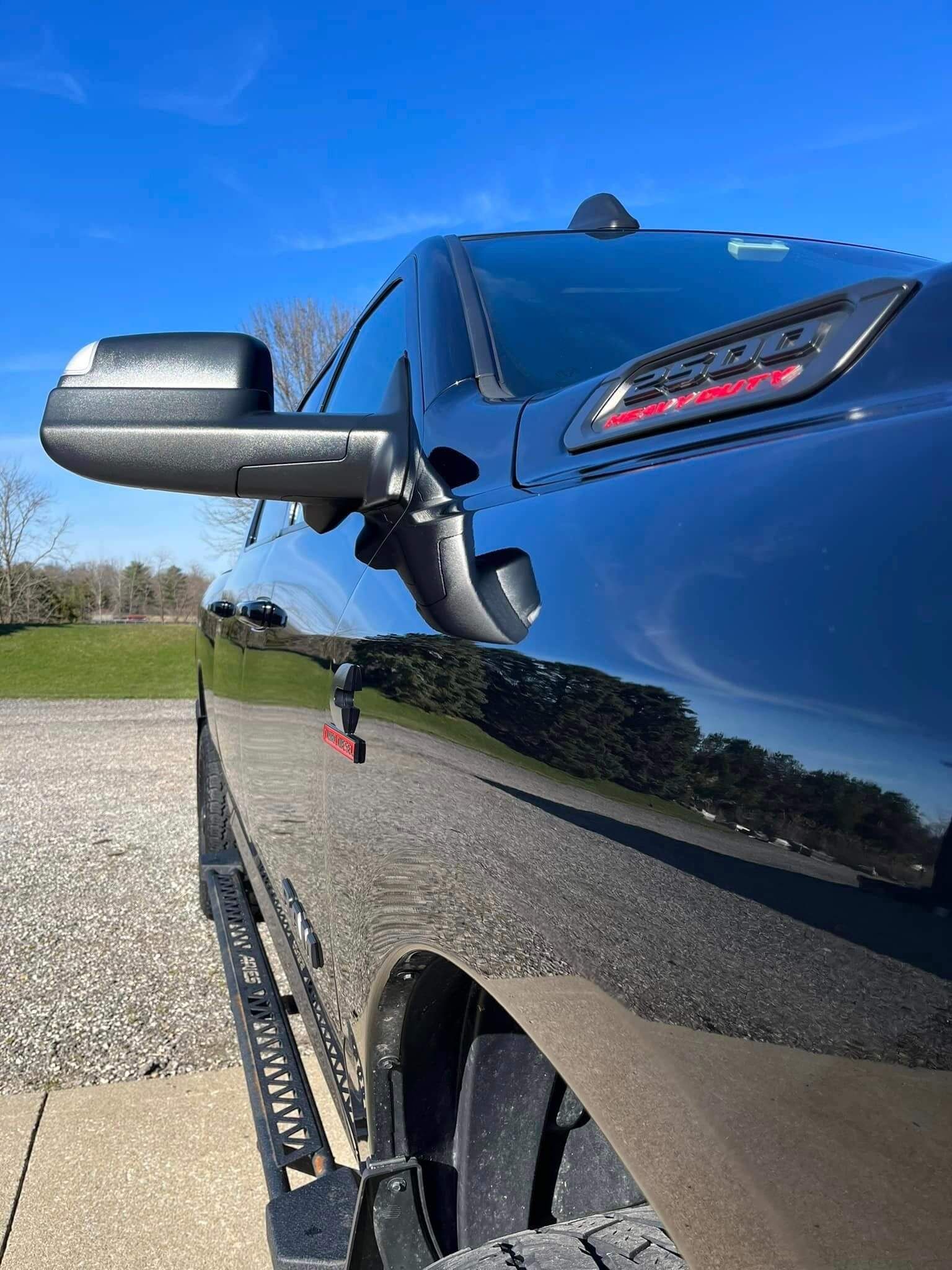 A side view of a glossy black Ram 1500 truck, featuring a side mirror, hood vent, and running board on a sunny, gravel lot.