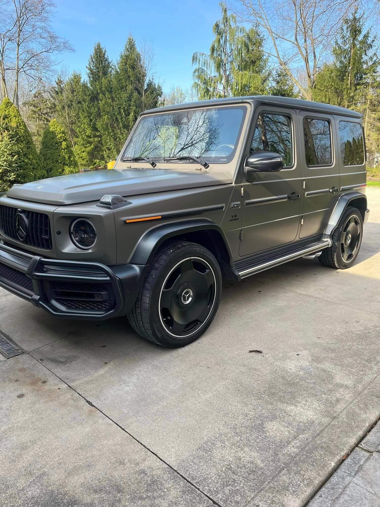 A matte olive-green Mercedes-Benz G-Wagon parked on a concrete driveway with trees in the background.