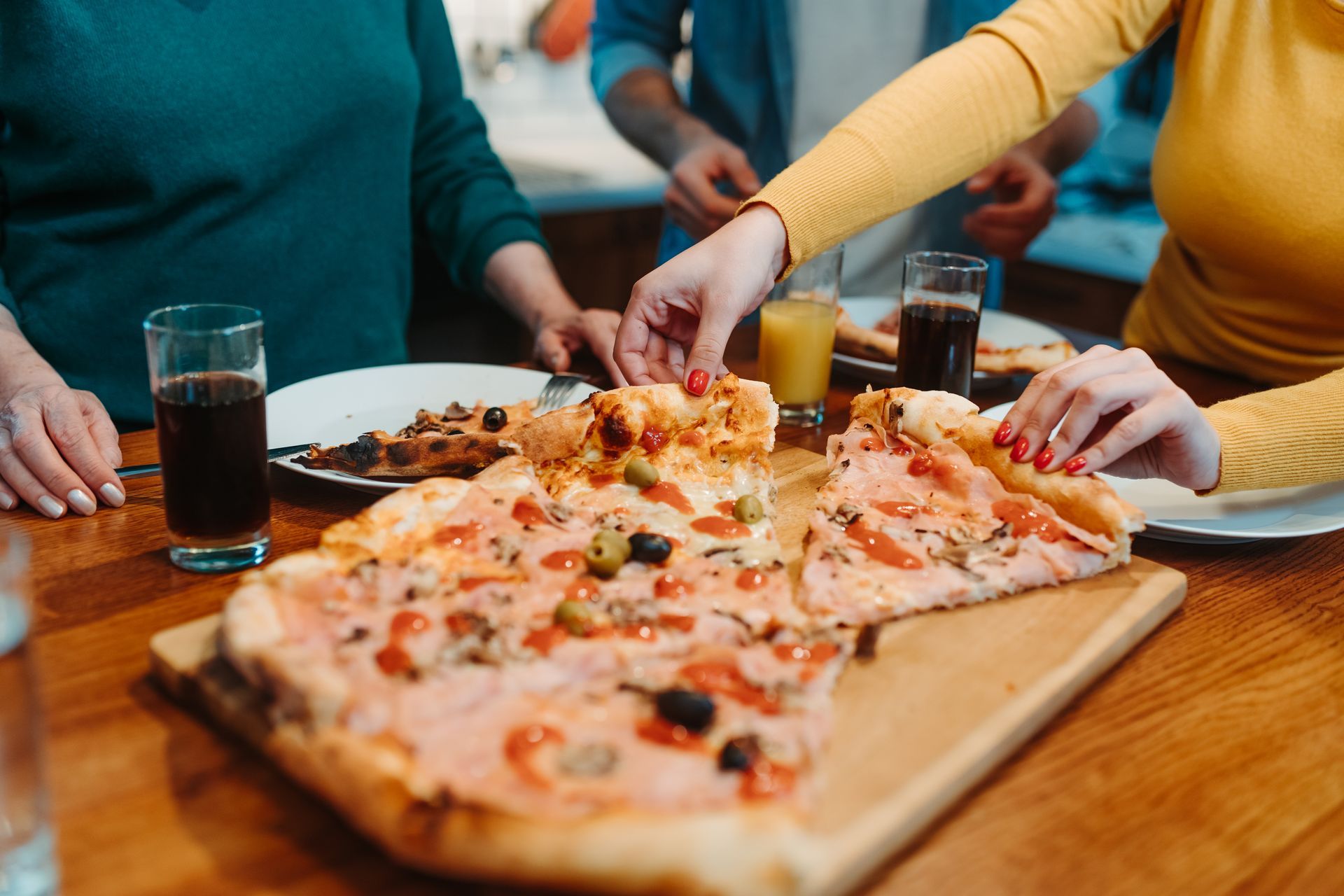 People reach for slices of pizza on a wooden board at a table, with drinks.