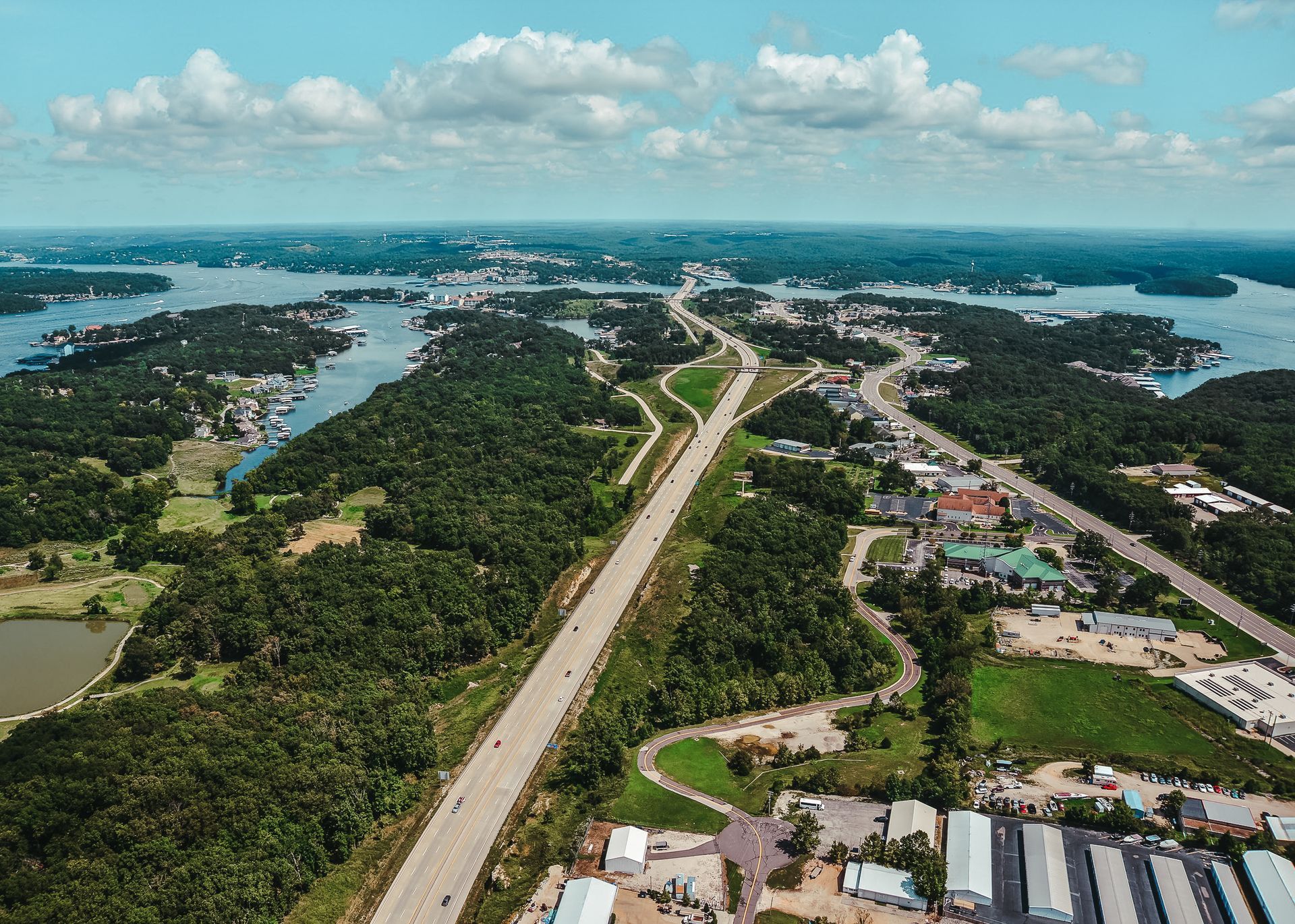 Aerial view of highway cutting through lush green forest, buildings, and a lake under a bright, cloudy sky.