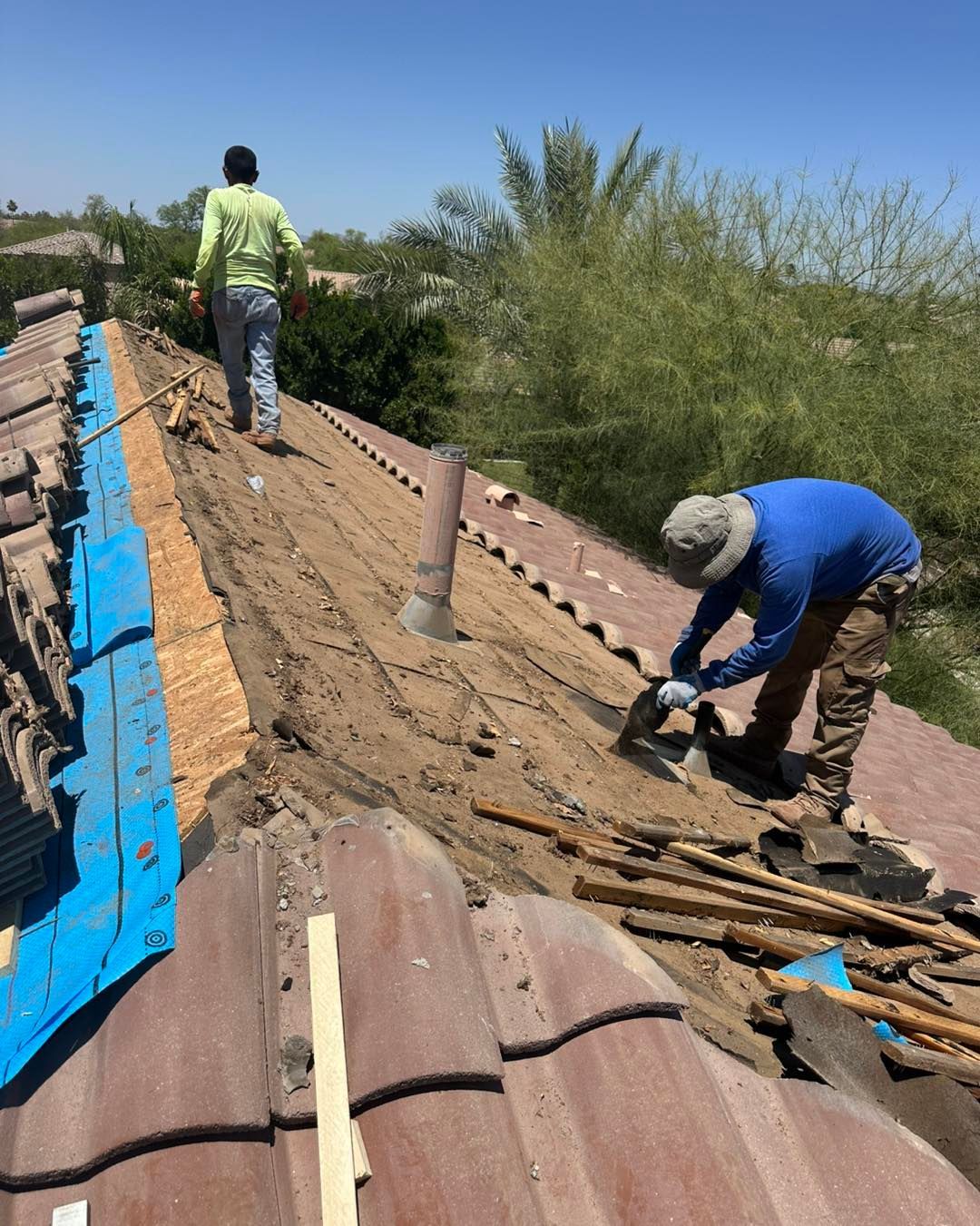 Two workers in bright shirts repair a residential roof with exposed wooden beams and tiles on a sunny day.