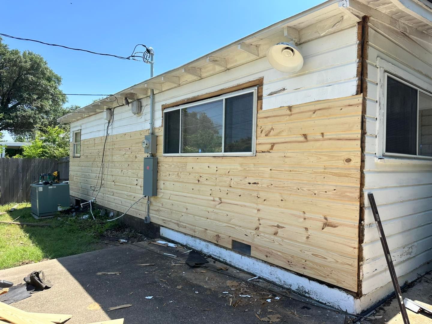 Exterior of a white house undergoing renovations, featuring a partially installed wood-paneled wall and new plywood sheathing.