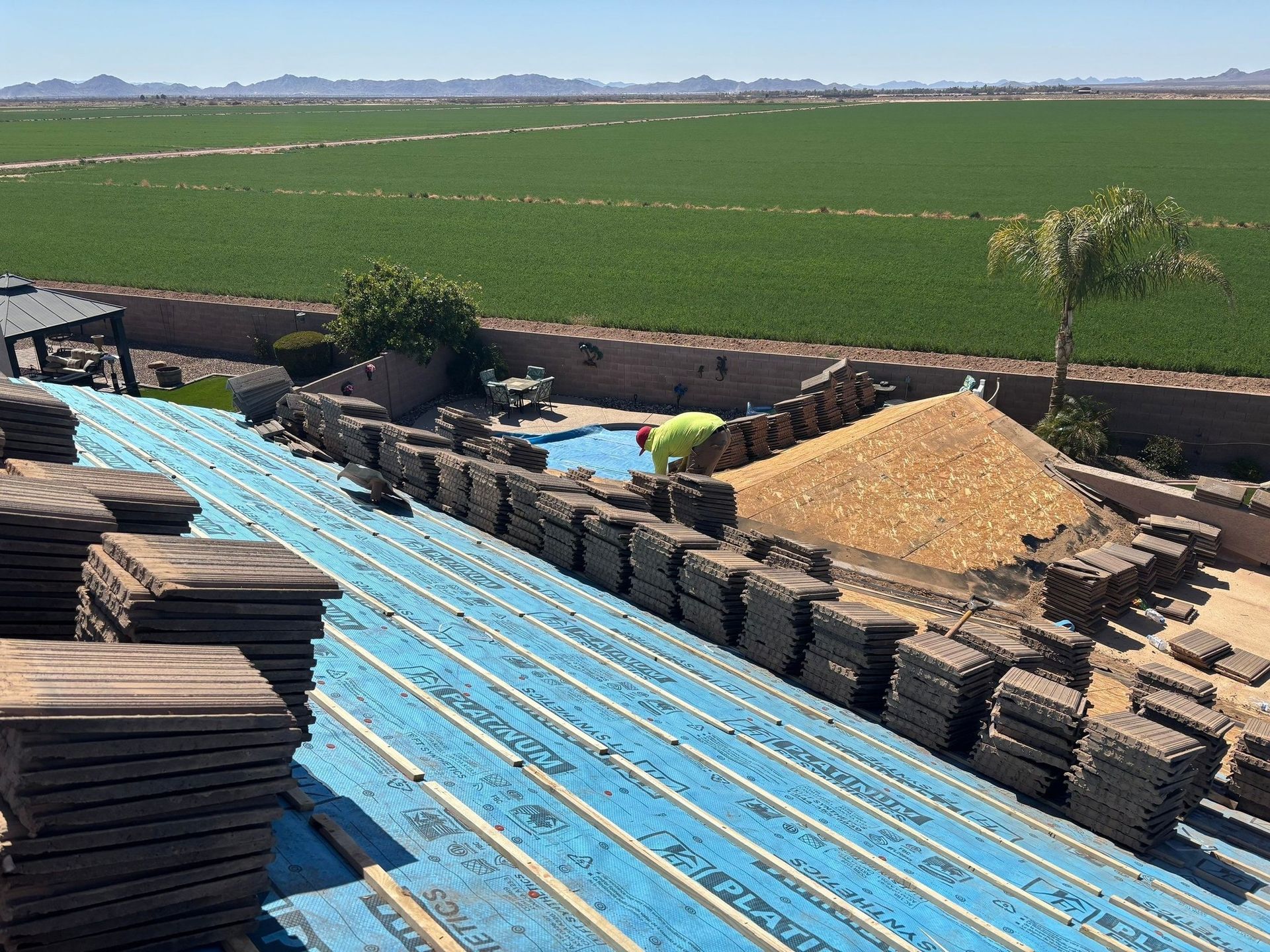 Workers install roof tiles on a house underlayment overlooking a sprawling field, with a pool and pile of gravel below.