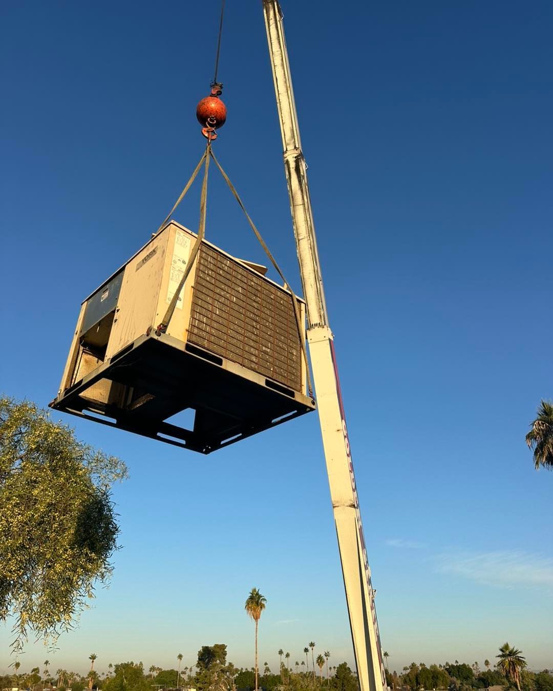 A crane lifts a large industrial HVAC unit through the air against a clear blue sky, with trees visible in the distance.