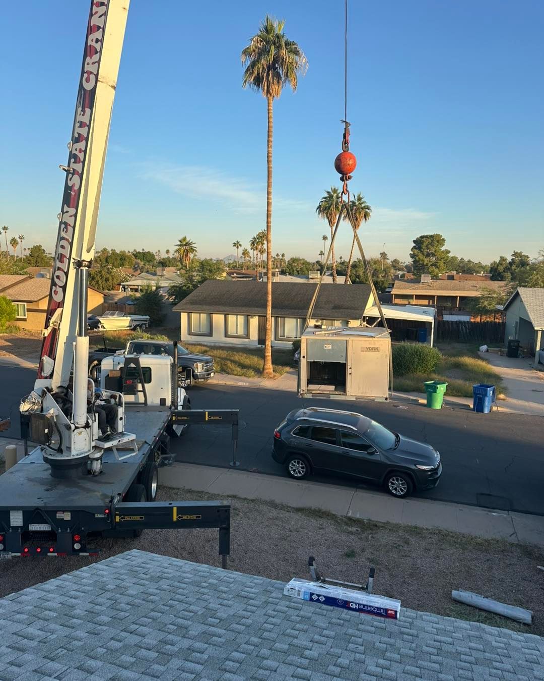A crane lifts a small structure over a residential street with parked cars and palm trees under a clear blue sky.