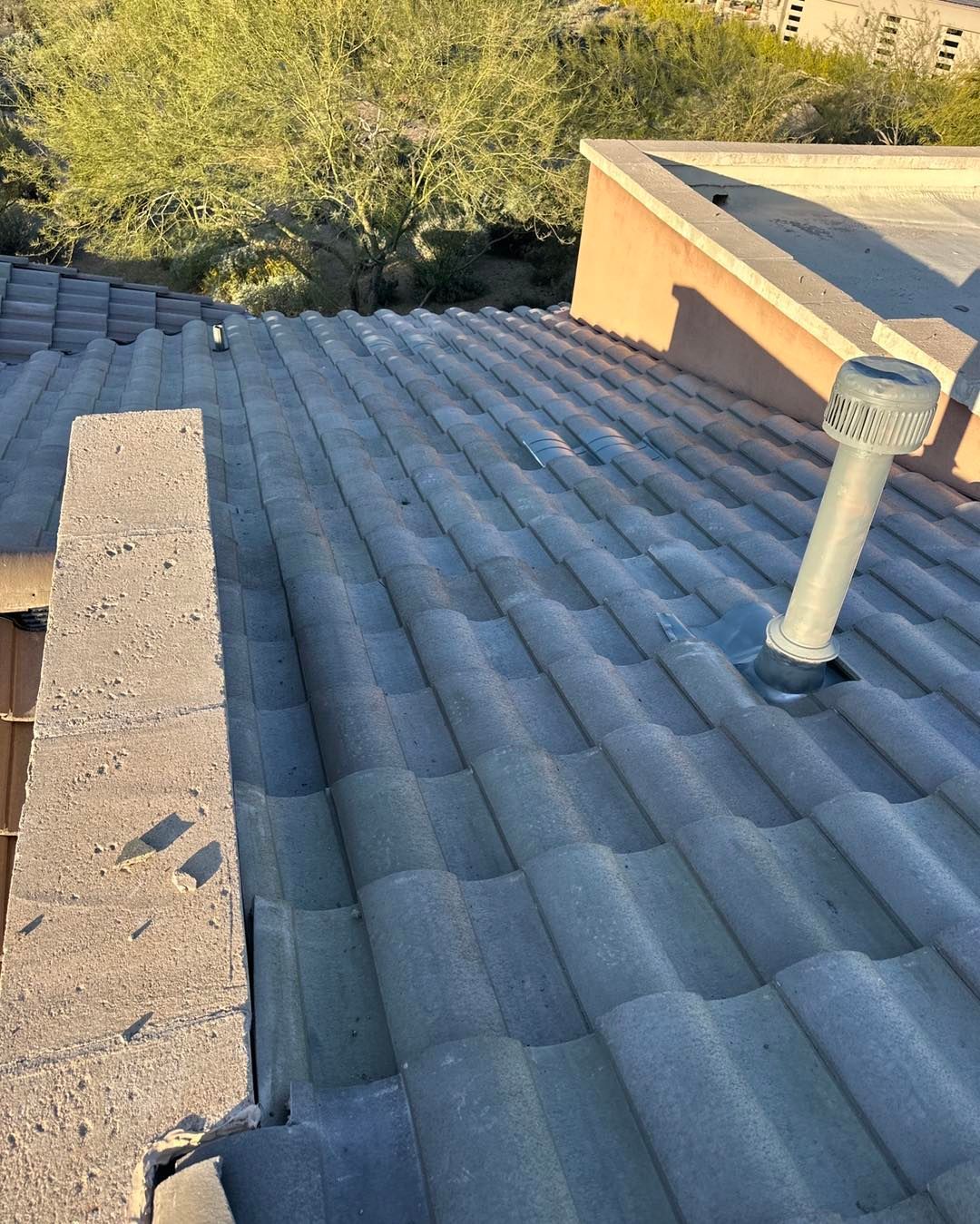 A gray tiled roof with a vertical vent pipe and part of a concrete chimney, overlooking a desert landscape with greenery.