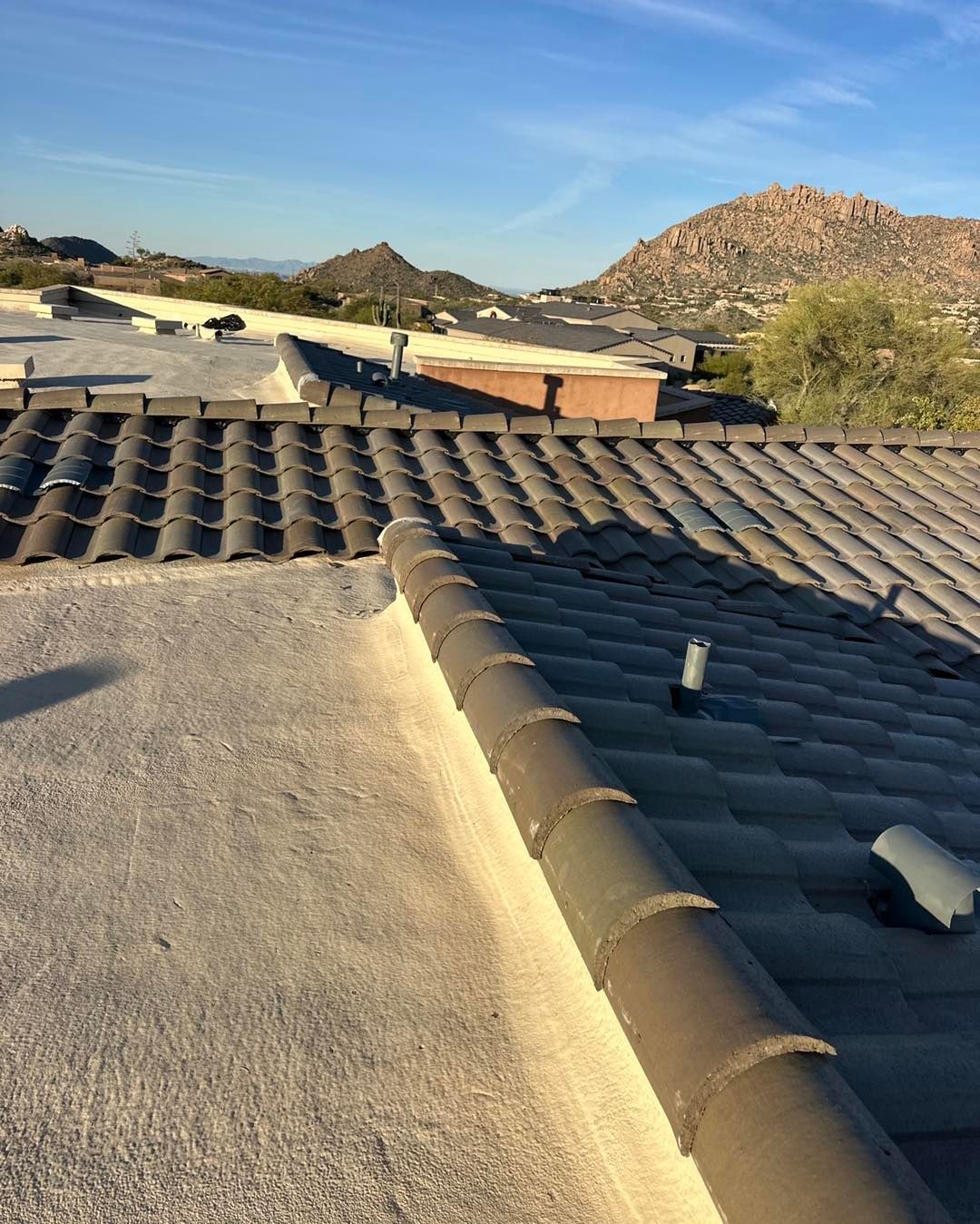A view from a roof showing grey tiles meeting a flat, light-colored section, with desert mountains under a clear blue sky.