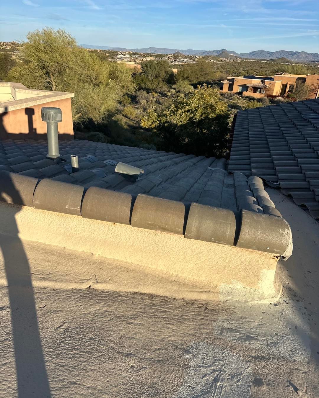 A rooftop view featuring a textured stucco parapet wall with dark cap tiles, overlooking a desert landscape with houses.