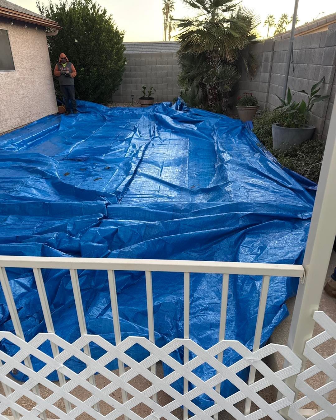 A blue tarp covers a swimming pool in a fenced backyard, with a person standing near the edge of the house.