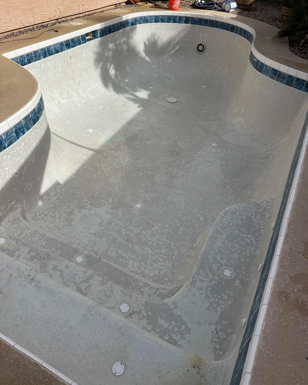 An empty, light-colored concrete swimming pool with blue decorative tile borders and a curved design.