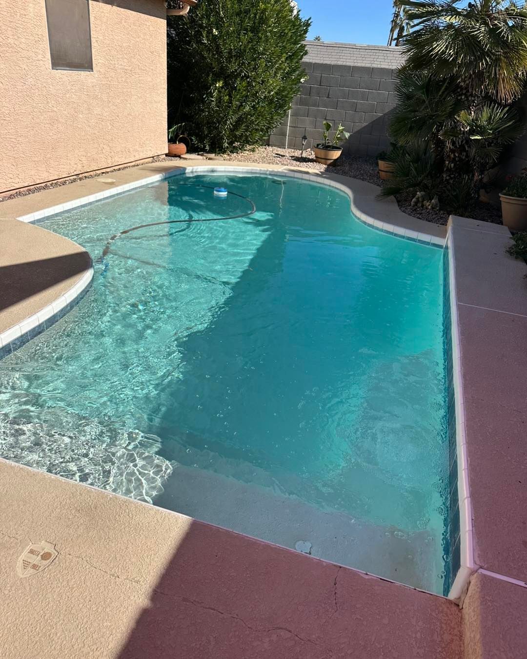 A turquoise swimming pool with integrated steps, set in a backyard patio next to a house wall and some greenery.