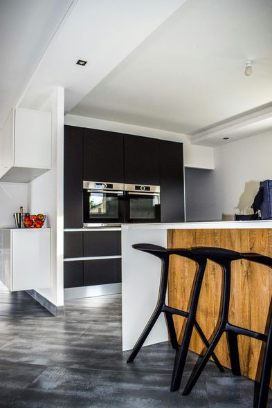 A kitchen island with two black stools faces dark cabinets and built-in ovens in a room with white walls and gray tile.