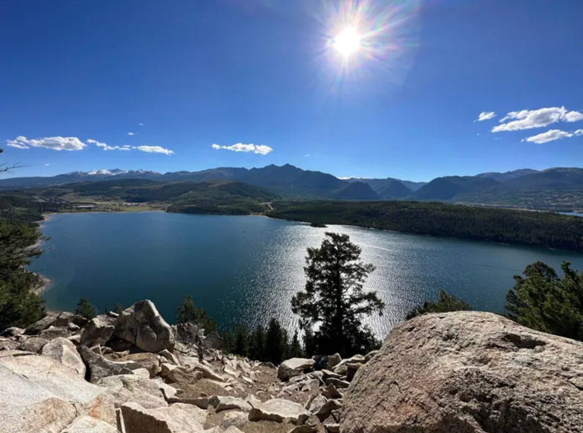 Vast lake shimmers under a bright sun, surrounded by mountains and trees. Rocky foreground. Blue sky.