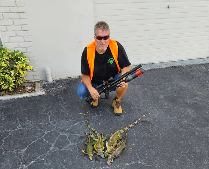 Man kneeling with rifle, surrounded by several iguanas on cracked pavement, near a garage and greenery.
