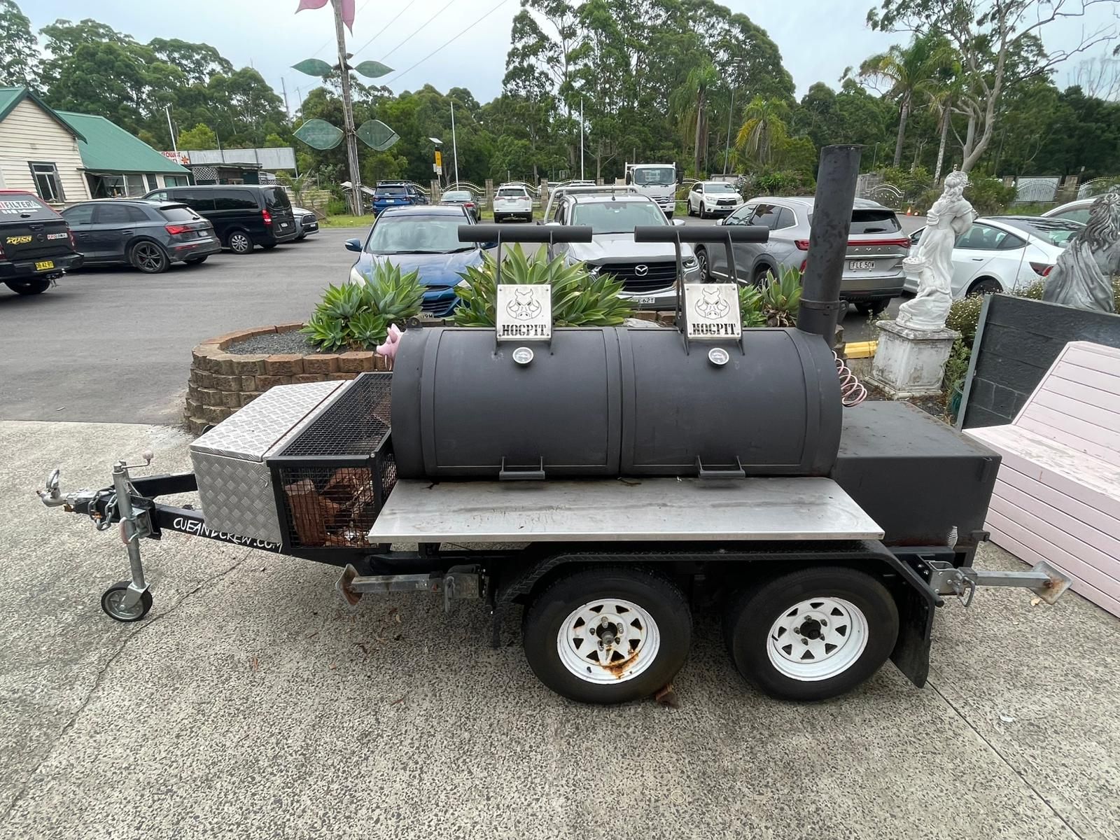 A black offset smoker trailer with two barrel chambers, a metal storage box, and a chimney, parked on a paved lot — Rustique on the Coast In Ourimbah, NSW