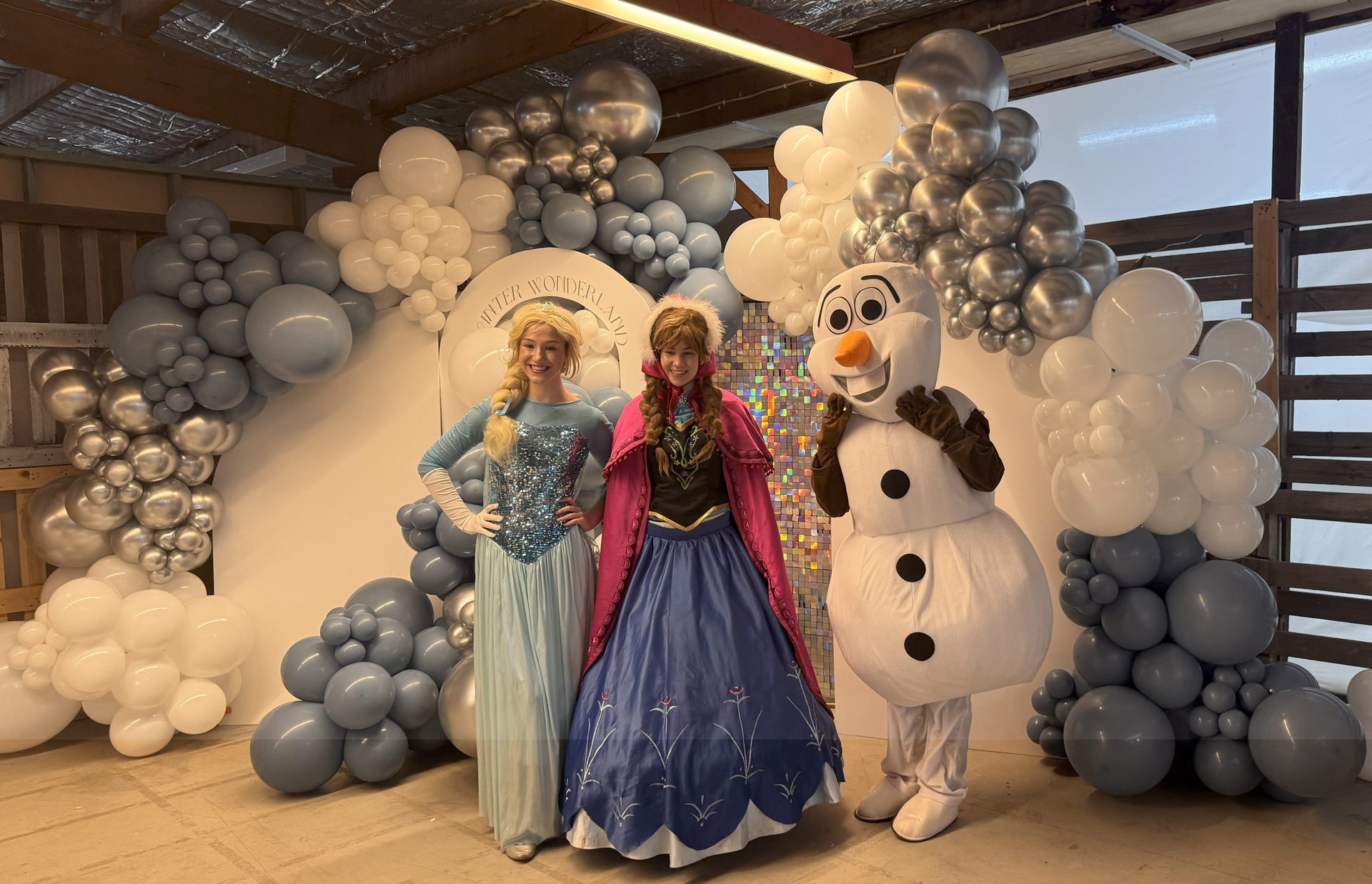 Elsa, Anna, and Olaf from Frozen pose in front of a blue and white balloon arch.