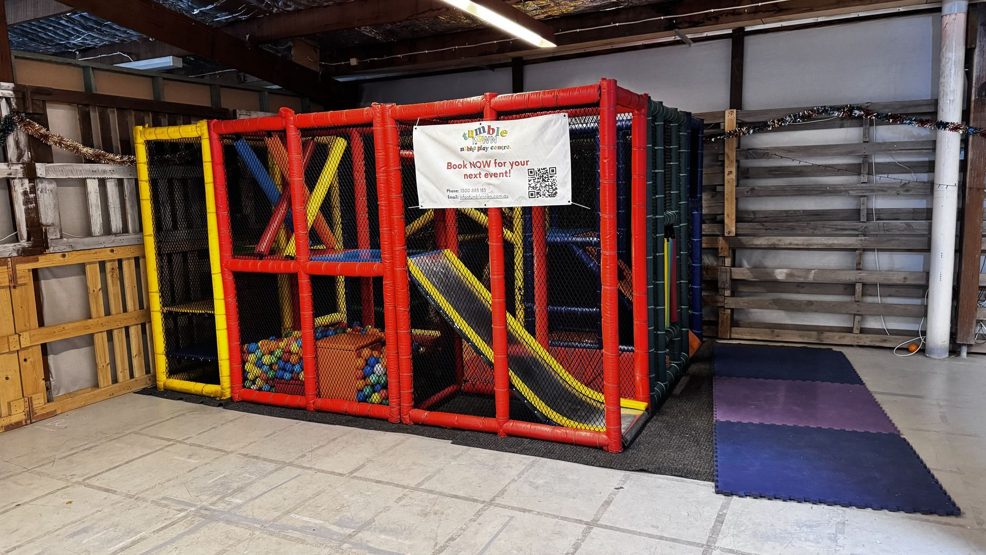 Indoor play structure with a red, yellow, and blue frame; a slide; and foam play mats on the floor.
