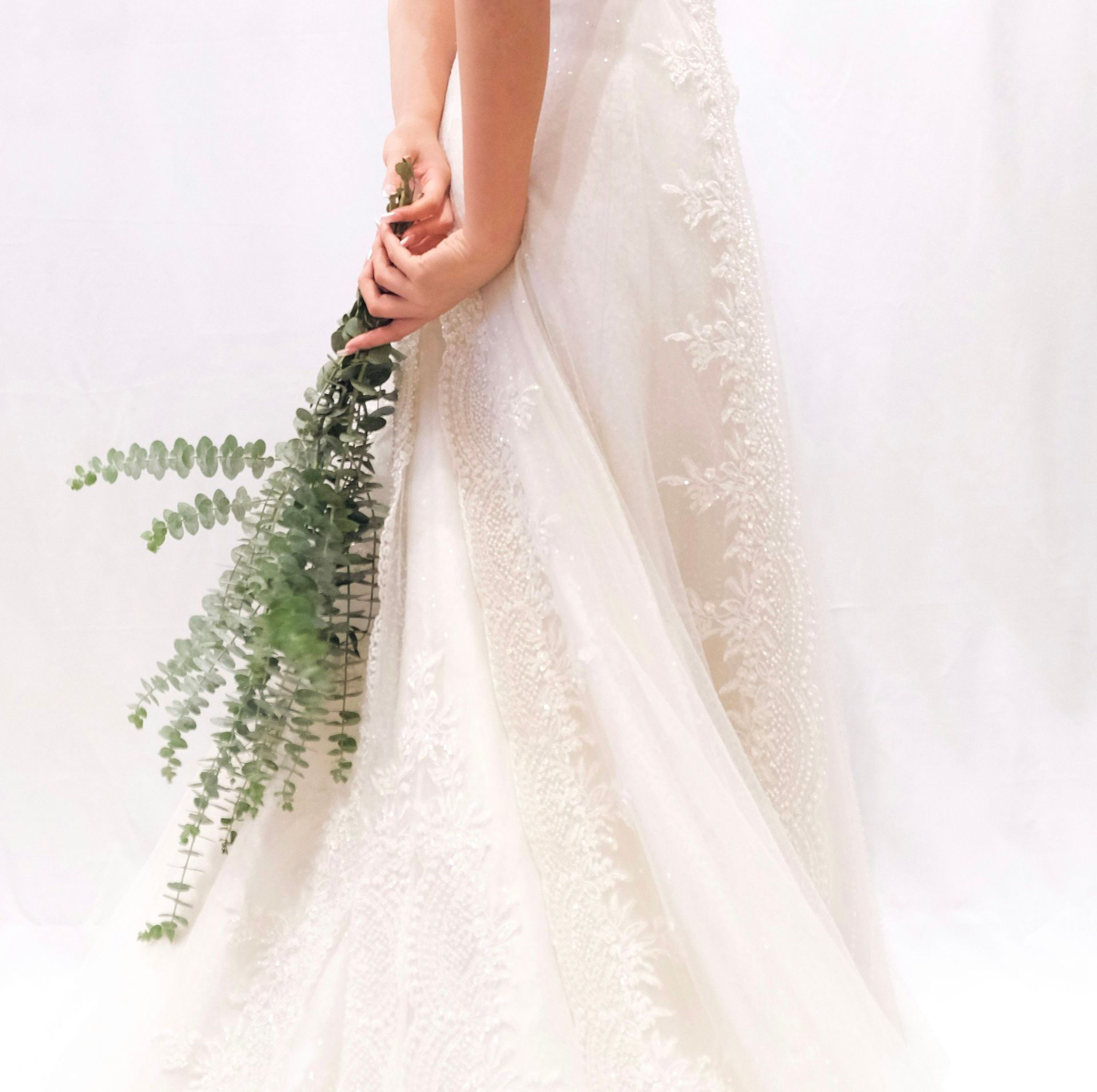 Woman in a white wedding dress holds a long, leafy green bouquet against her back, standing in front of a white background — Rustique on the Coast In Ourimbah, NSW