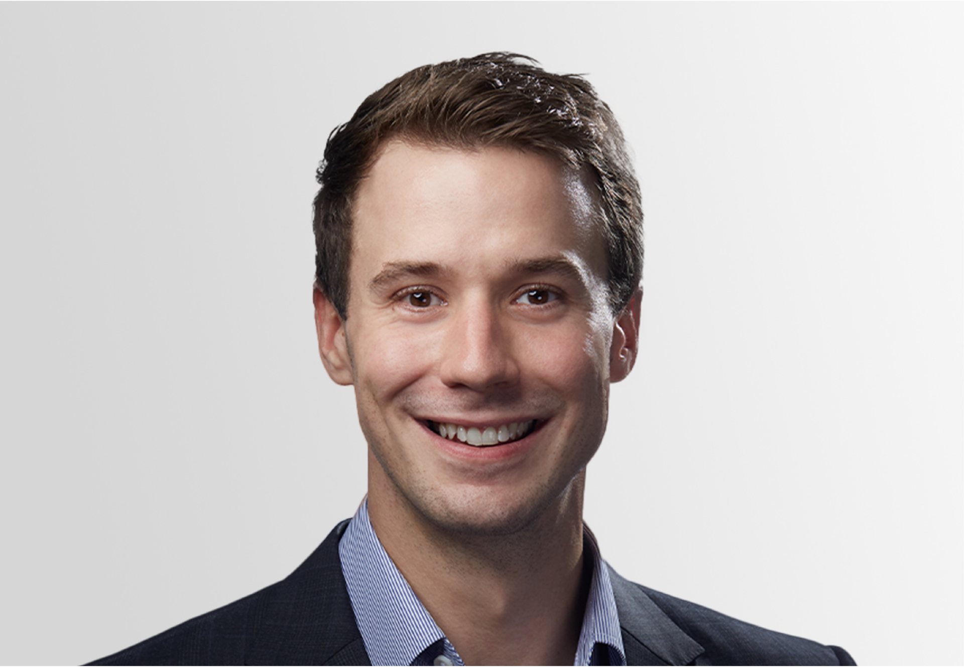 Man smiling, wearing a blue pinstripe shirt and dark blazer, against a light gray background.