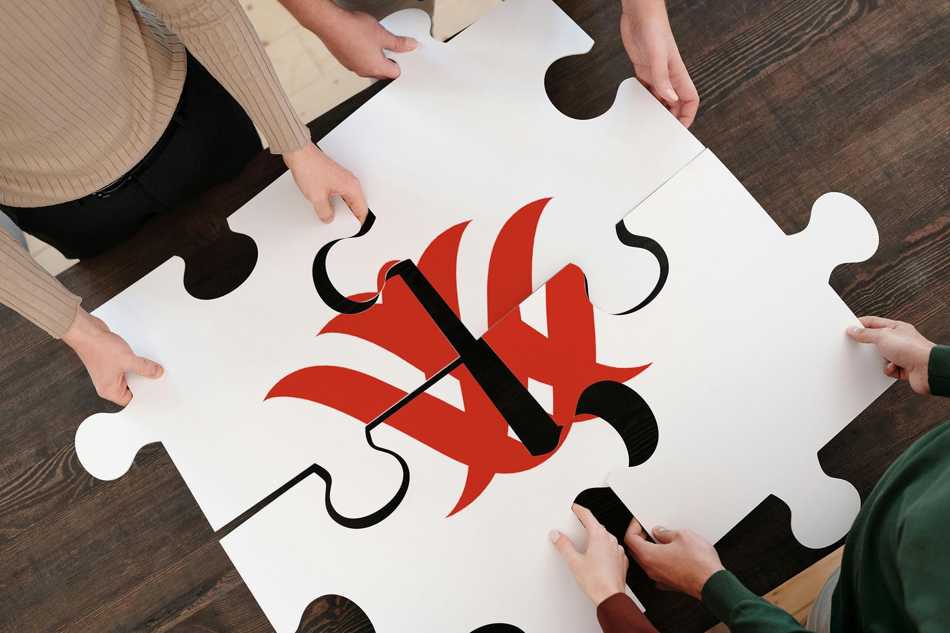 Hands assemble giant puzzle with red emblem on wood floor.