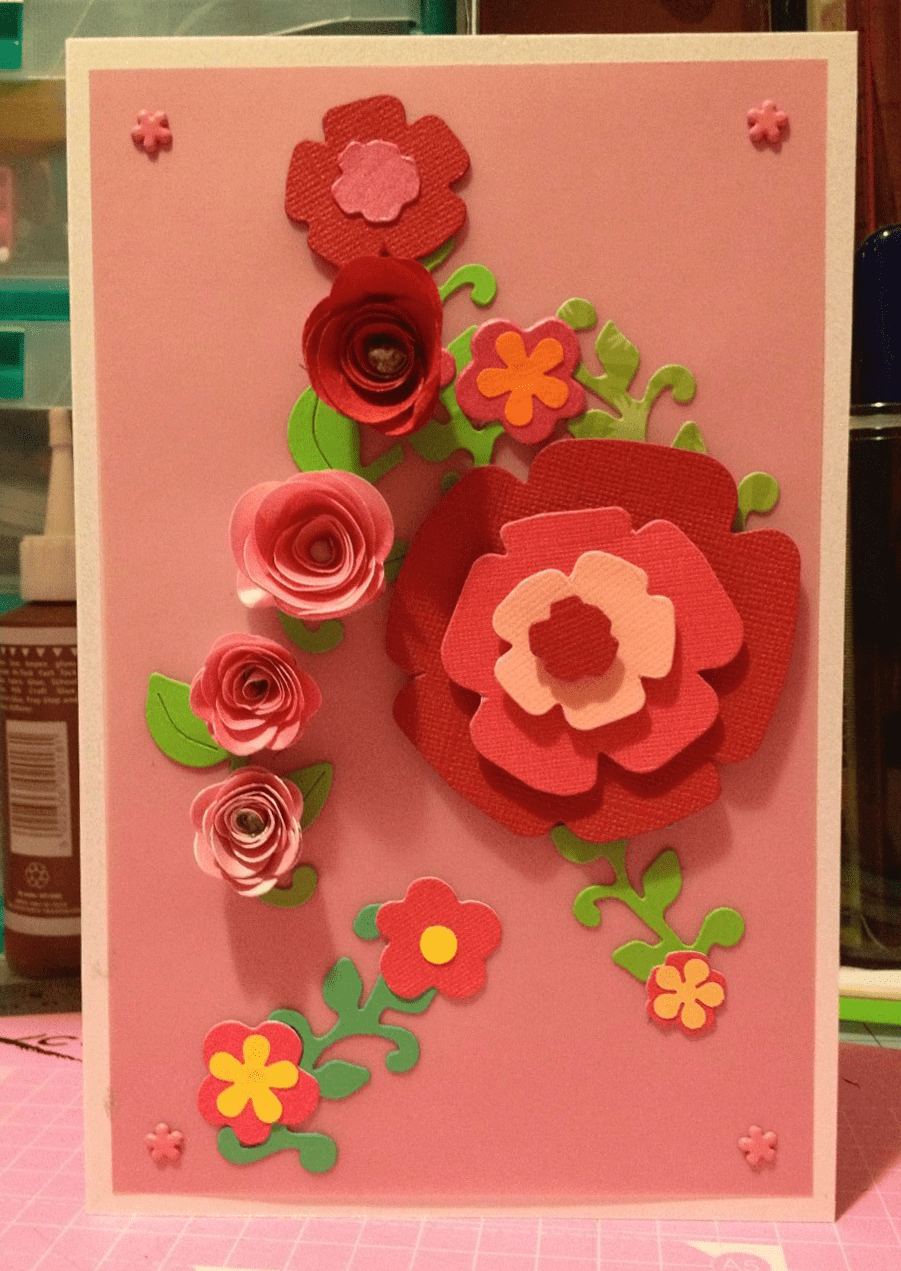 A side-folded A5 card with a pink paper cover displaying a cascade of small pink and red handmade paper flowers surrounding a larger, single pink and red handmade paper bloom, all with leafy greenery.