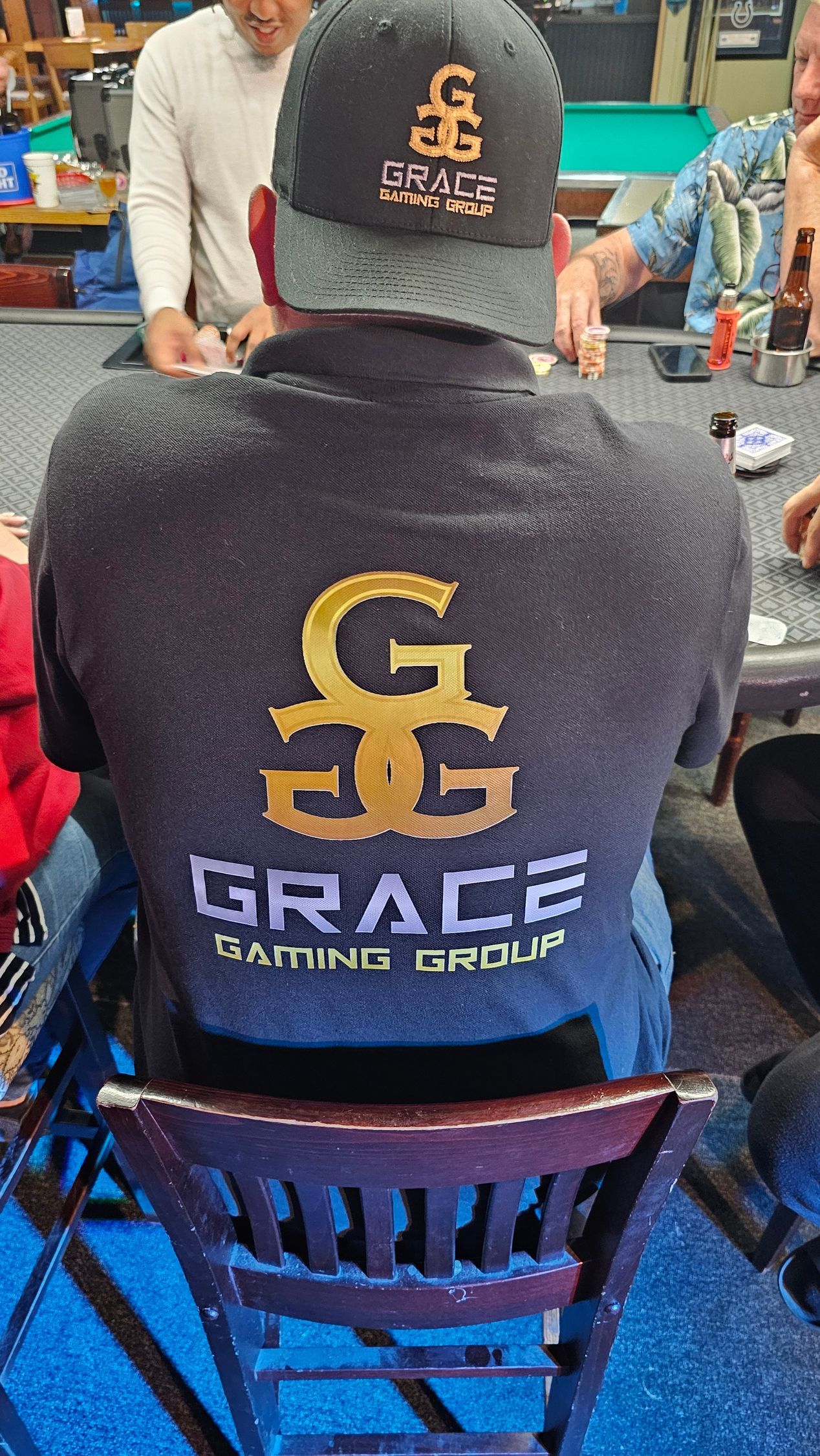 A man wearing a grace gaming group shirt is sitting in a chair.