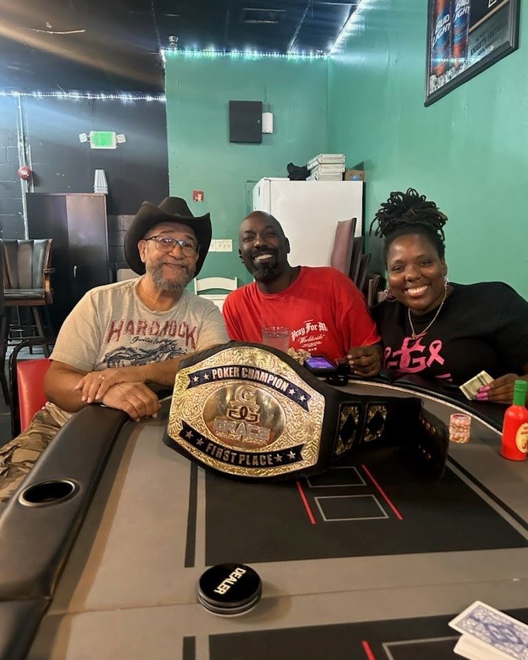 Three people smiling, holding a poker championship belt at a table in a bar.