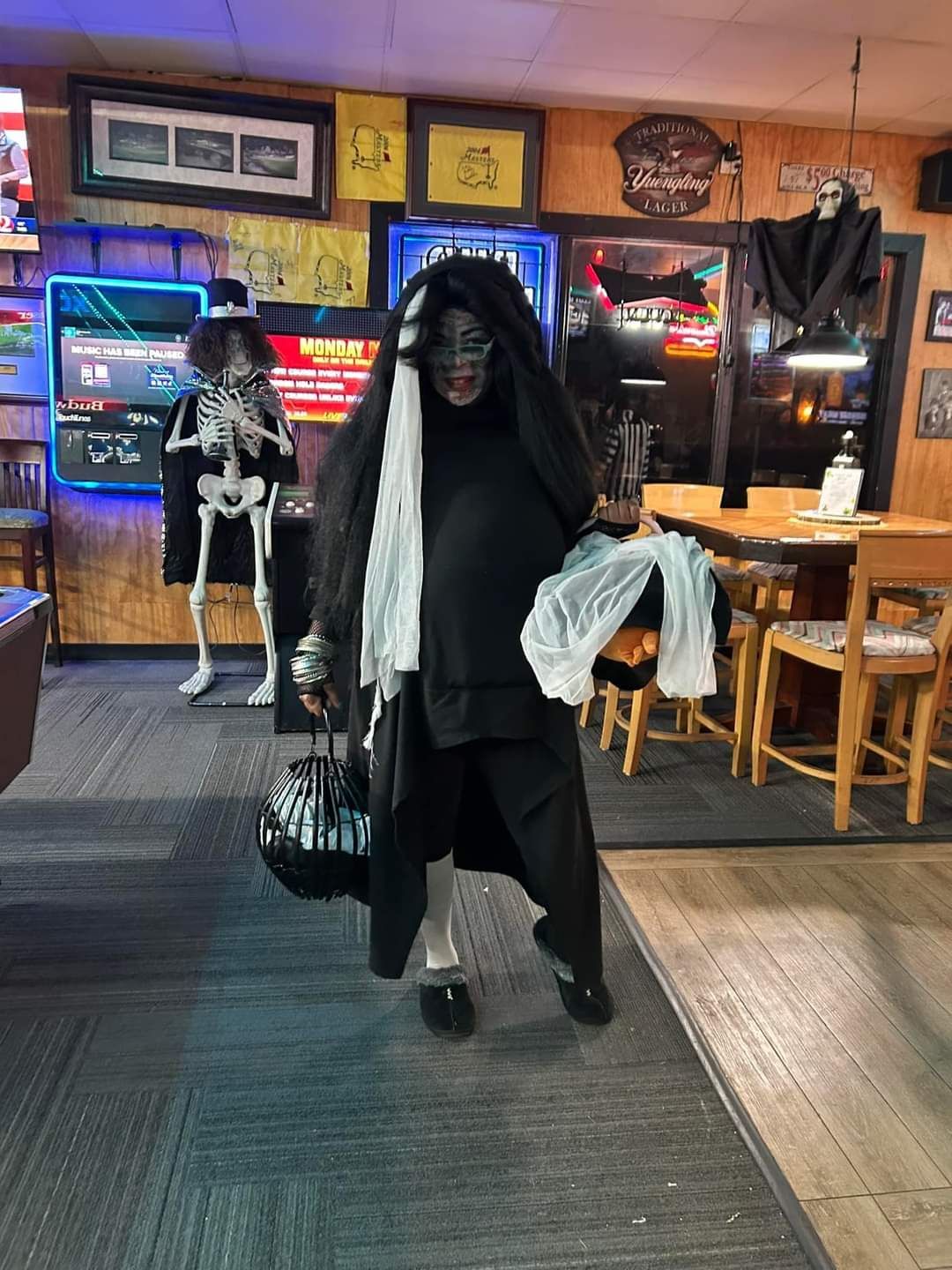 A woman in a black costume is standing in a room with a skeleton in the background.