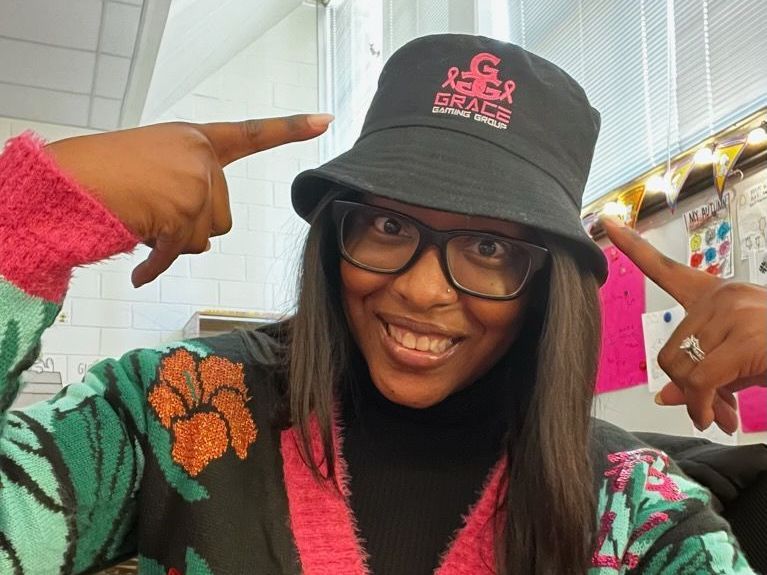 A woman wearing a bucket hat and glasses is pointing at herself.