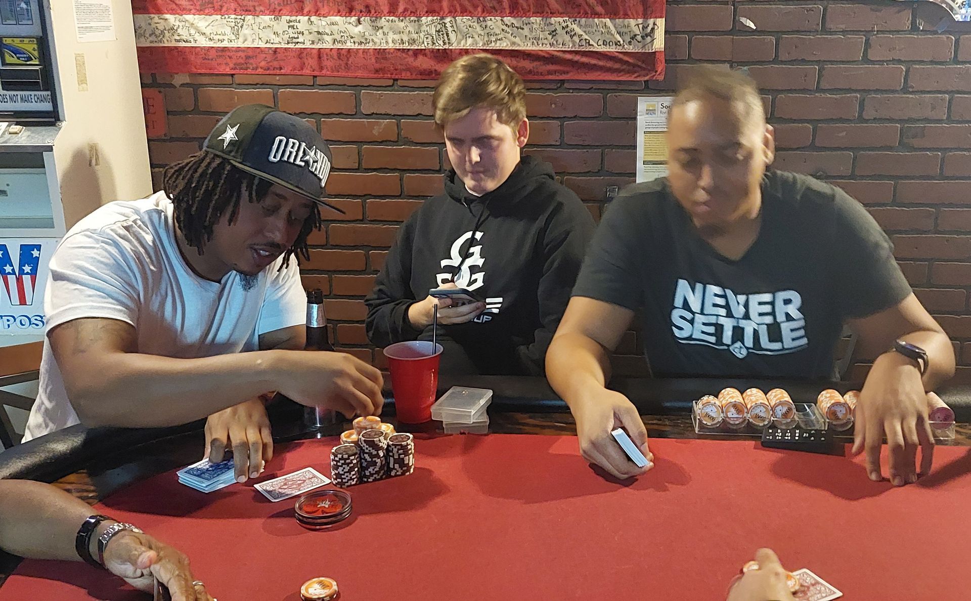 Three men are playing poker at a table and one of them is wearing a shirt that says never settle.