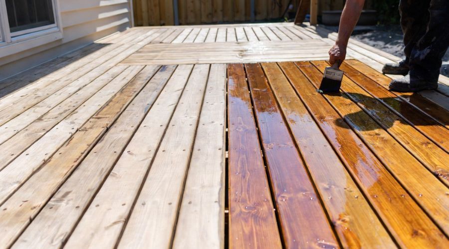 A person uses a brush to apply a warm-toned stain to a wooden deck, showing a clear contrast between raw and finished wood.